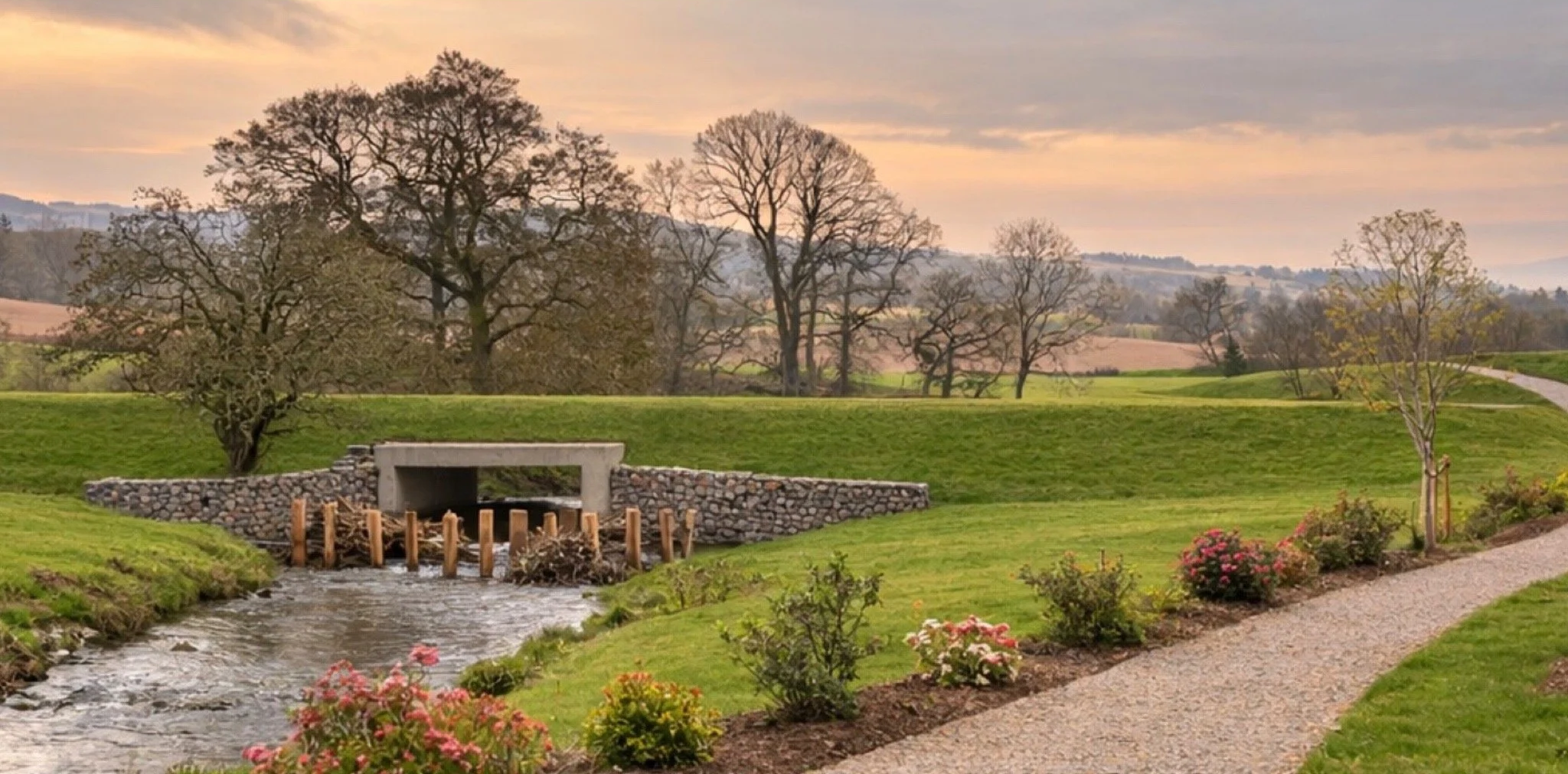 A scenic landscape with a small stream flowing under a concrete bridge, surrounded by lush green grass, budding trees, and a flower bed. The background shows rolling hills and a cloudy sky at sunset.