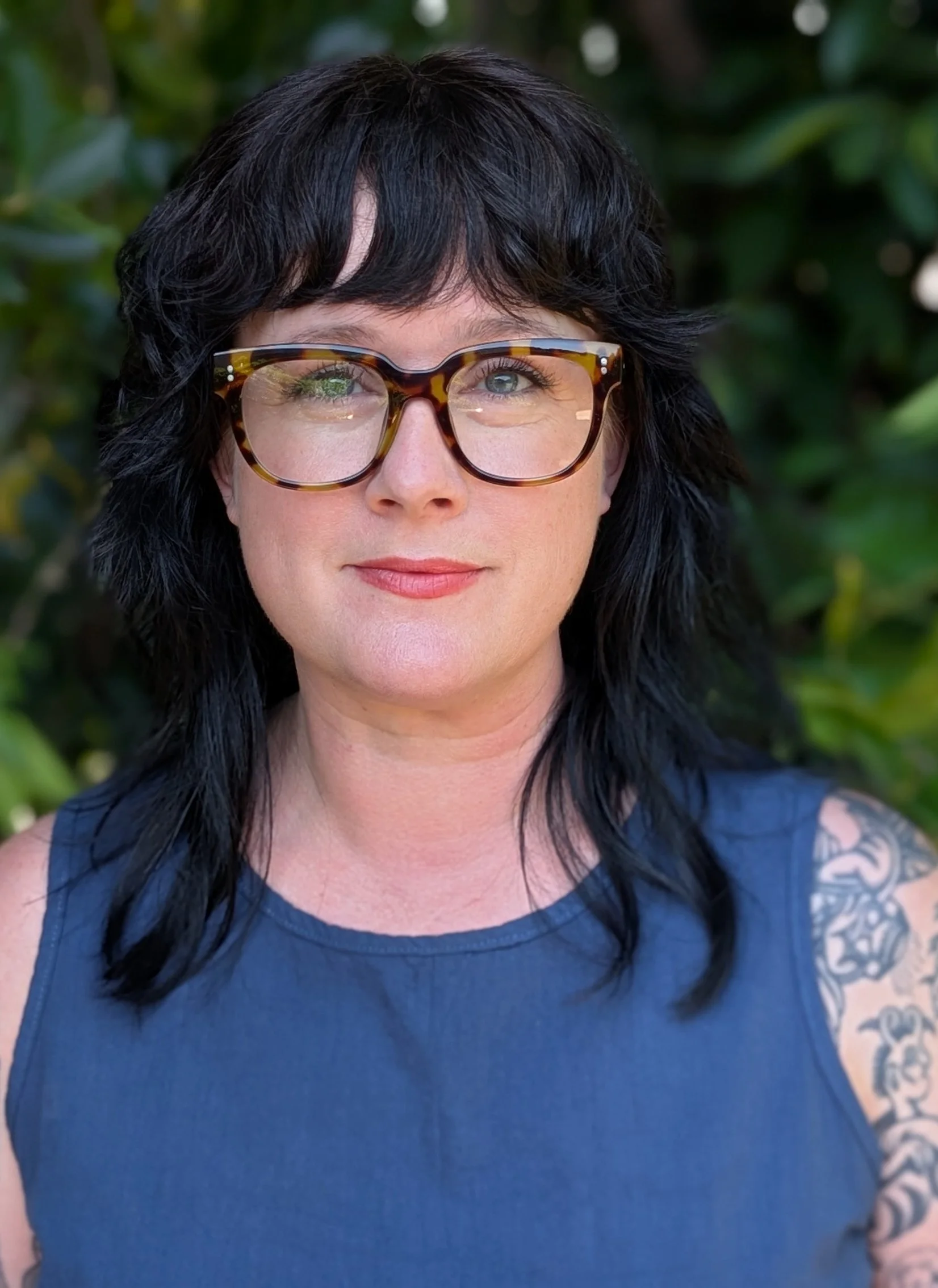 Jenny Walton-Wetzel. A headshot of Jenny Walton-Wetzel, A woman with black curly hair, wearing tortoiseshell glasses and a navy blue sleeveless top, smiling outdoors with green foliage in the background.