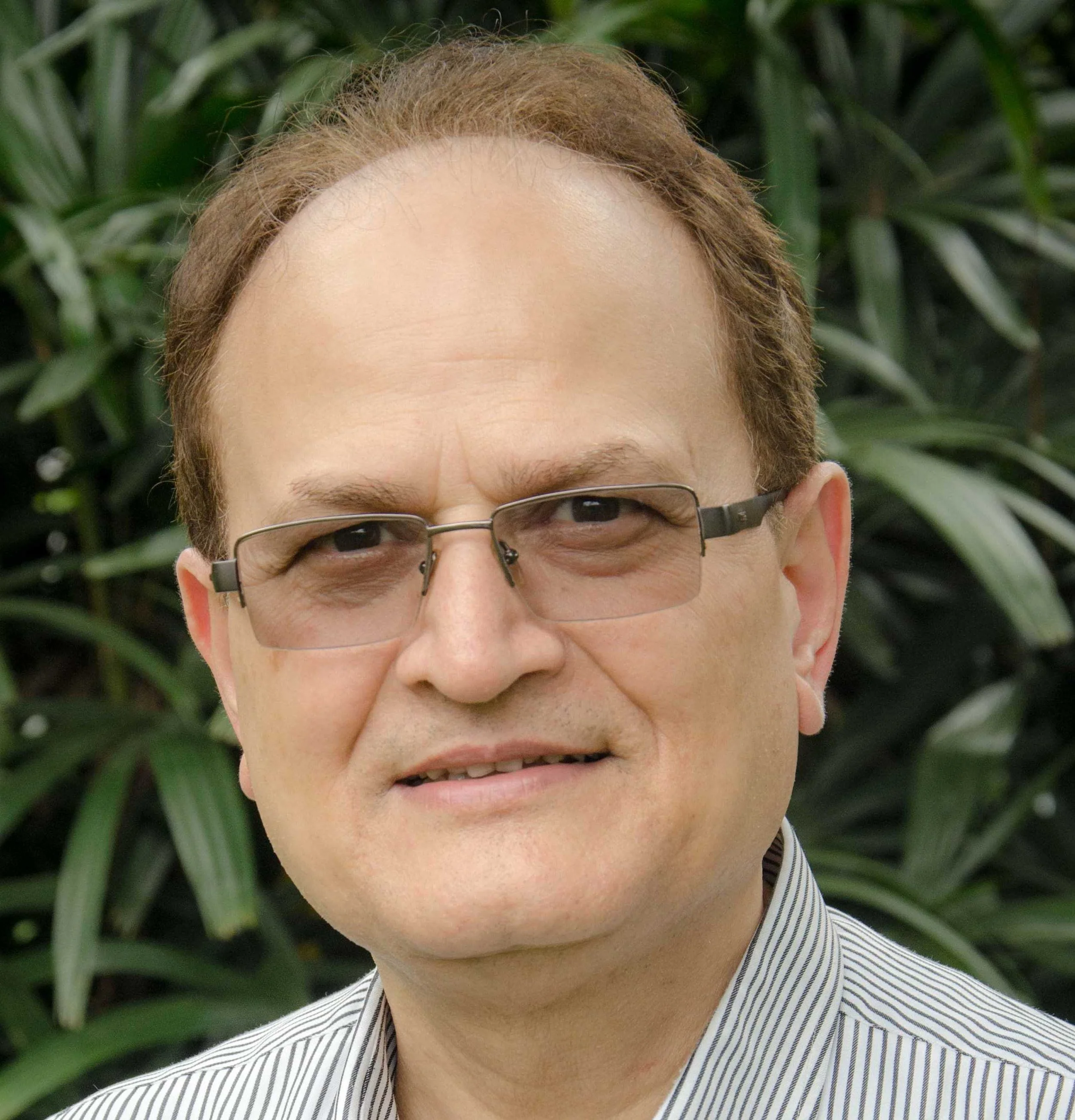 Close-up of a man with light skin, short reddish hair, wearing glasses, and a striped shirt, outdoors with green foliage in the background.