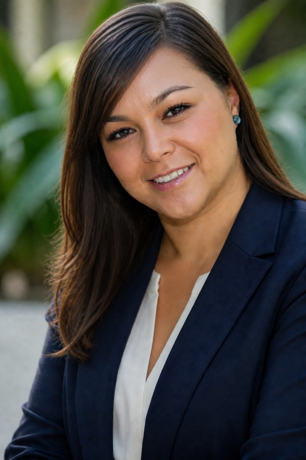 A woman with long dark hair wearing a navy blazer and white blouse, smiling outdoors with blurred green foliage in the background.