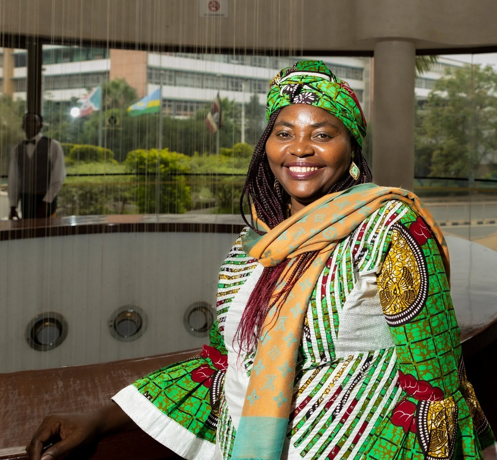 Smiling woman wearing vibrant green, yellow, and red clothing and a headwrap, sitting in an indoor space with large glass windows showing a building, flags, and landscaped greenery outside.