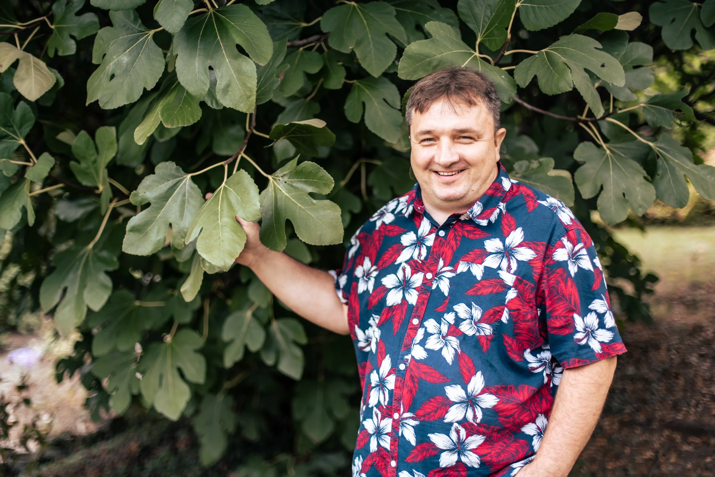 A smiling man in a colorful Hawaiian shirt standing outdoors surrounded by large green leaves.