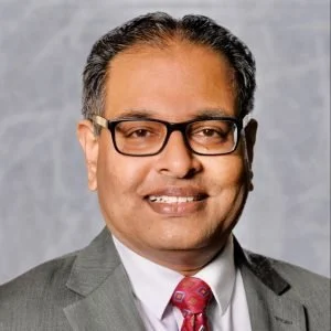 Headshot of a man in a suit with glasses, smiling, against a gray background.