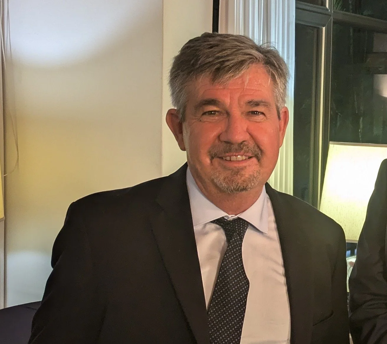 A middle-aged man with gray hair and a beard, wearing a black suit, white shirt, and dark dotted tie, smiling indoors near a window with green curtains and a lit lamp.