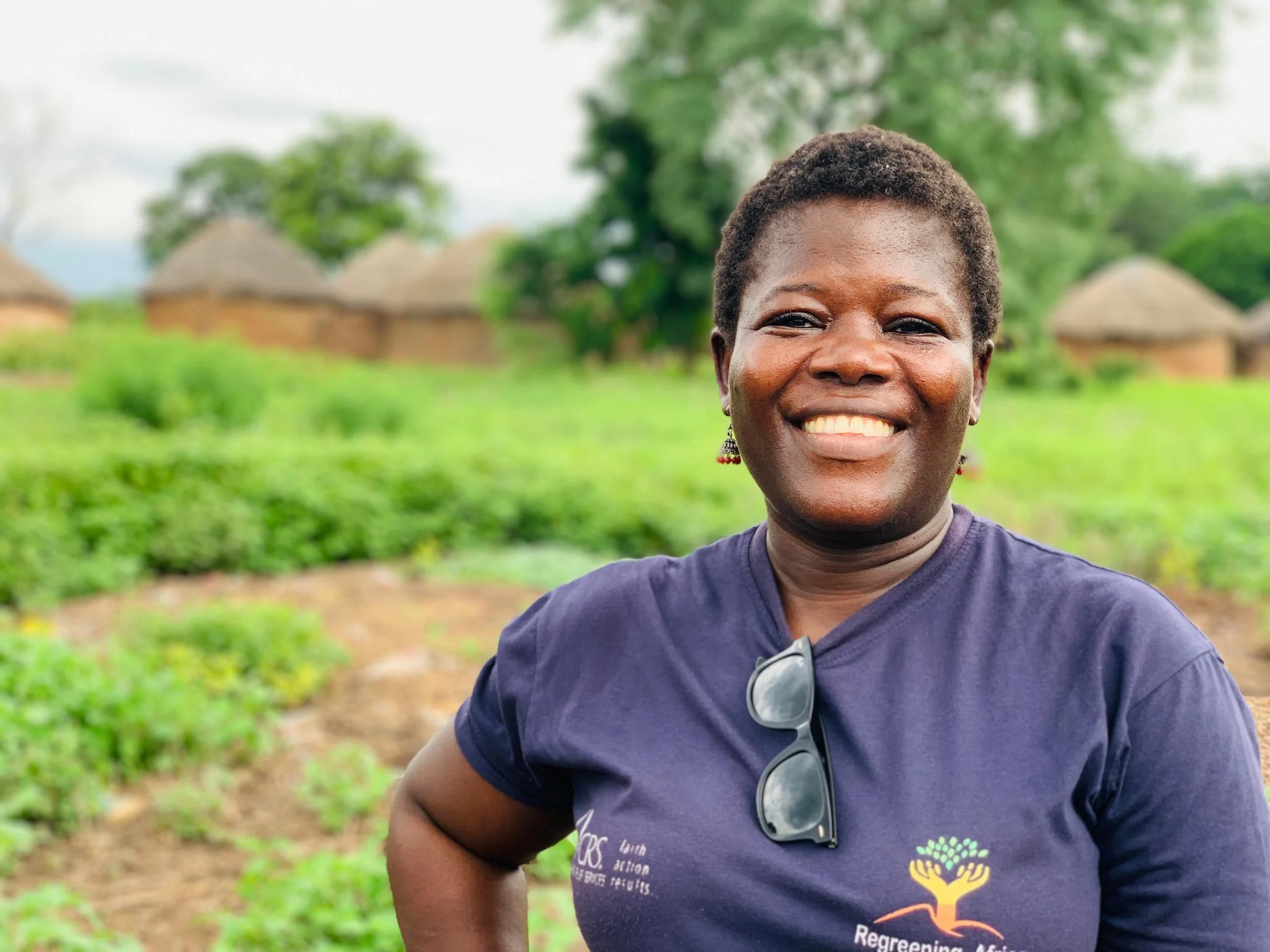 A smiling woman wearing a navy blue shirt and glasses hanging around her neck. She is standing outdoors in a rural setting with small thatched-roof huts and green trees in the background.