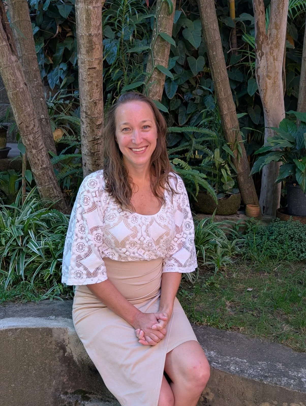 A woman with light skin and brown hair sitting on a stone bench in a lush garden with trees and green plants, smiling at the camera.