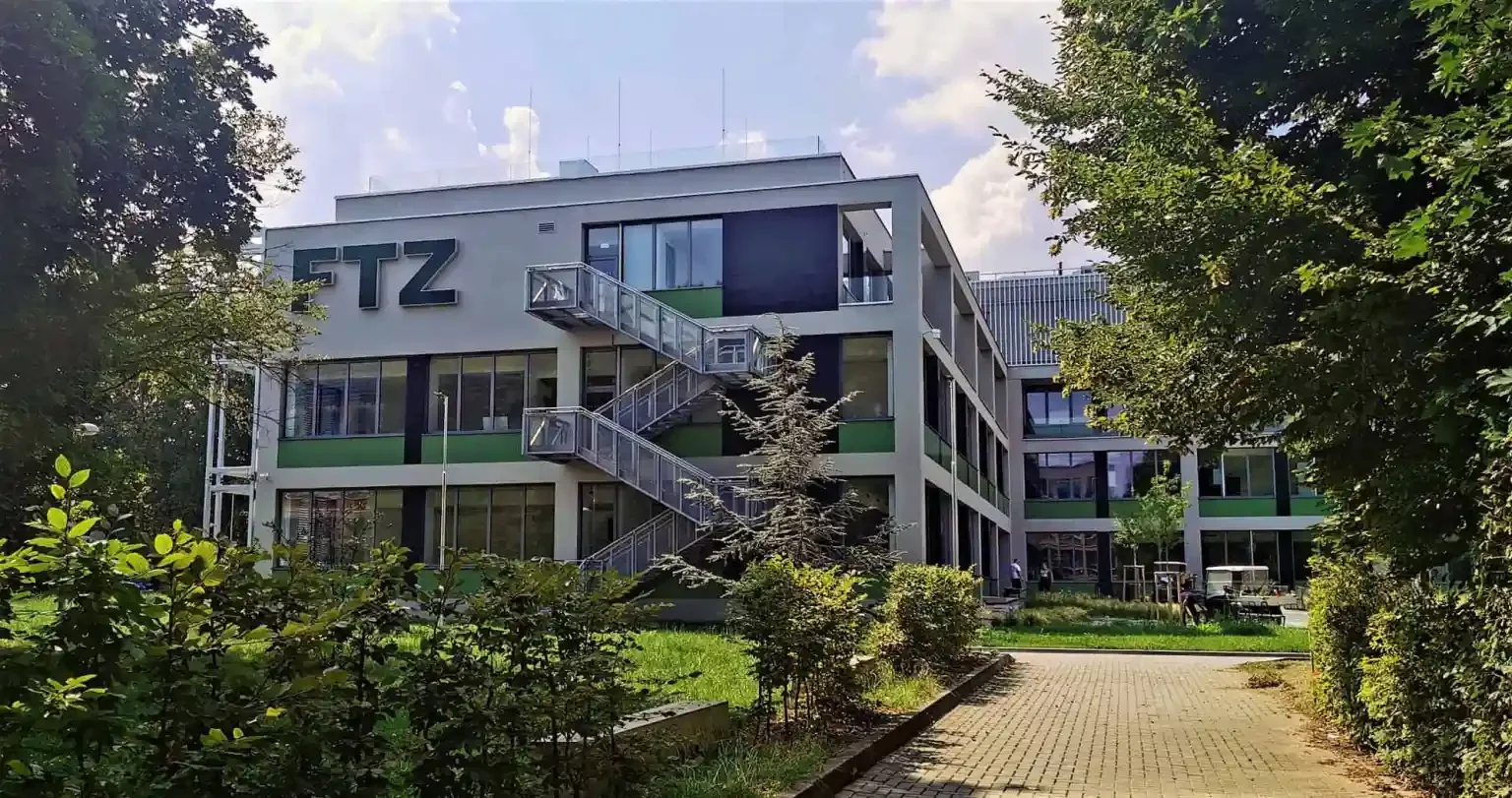Modern four-story office building with large windows, outdoor staircase, and the letters 'ETZ' on the top left corner. Surrounded by trees, bushes, and a paved walkway.