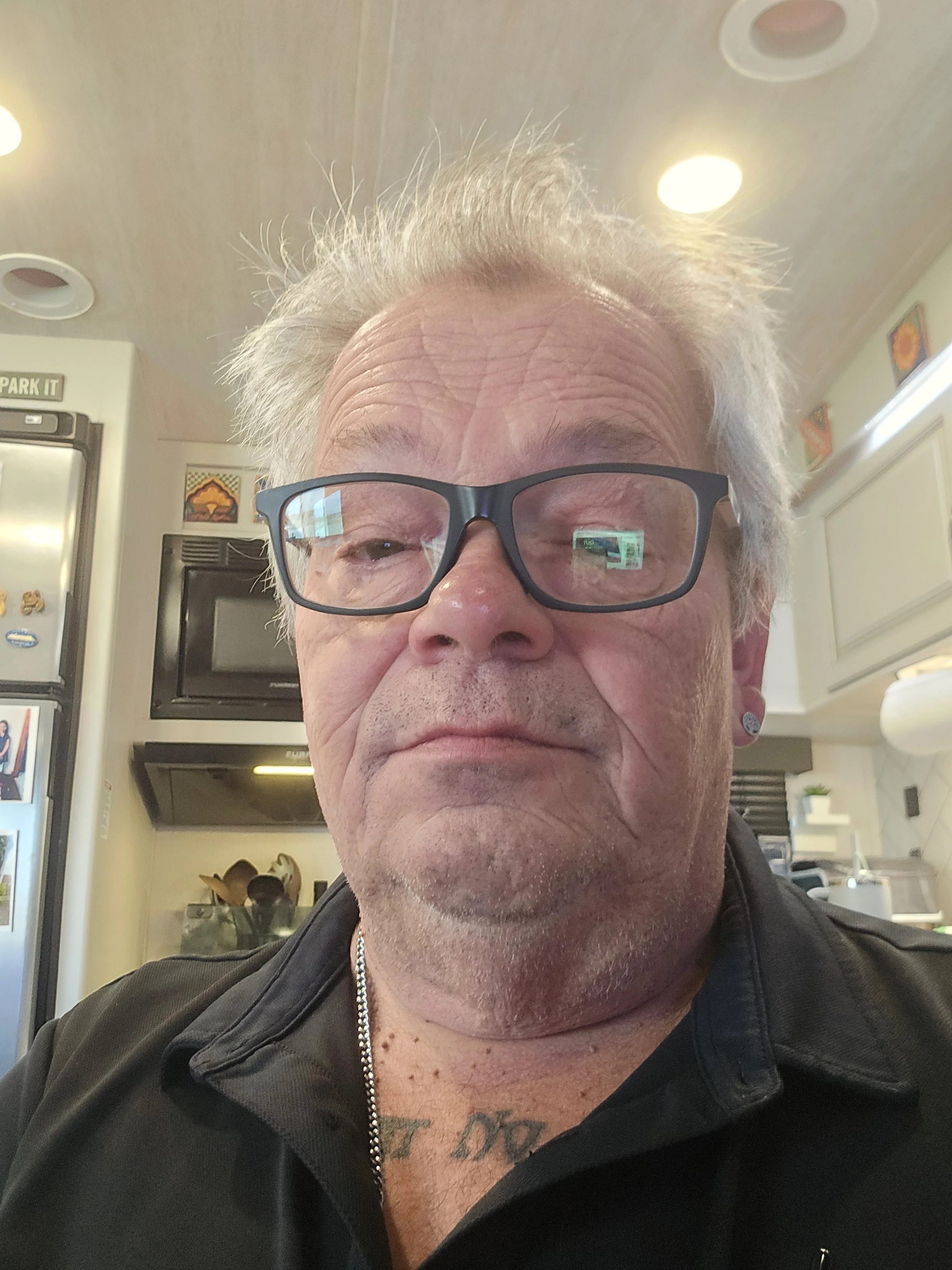 Close-up of an older man with glasses, gray hair, and tattoos on his chest and neck, wearing a black leather jacket, in a room with white cabinets, a TV, and decorative wall art.