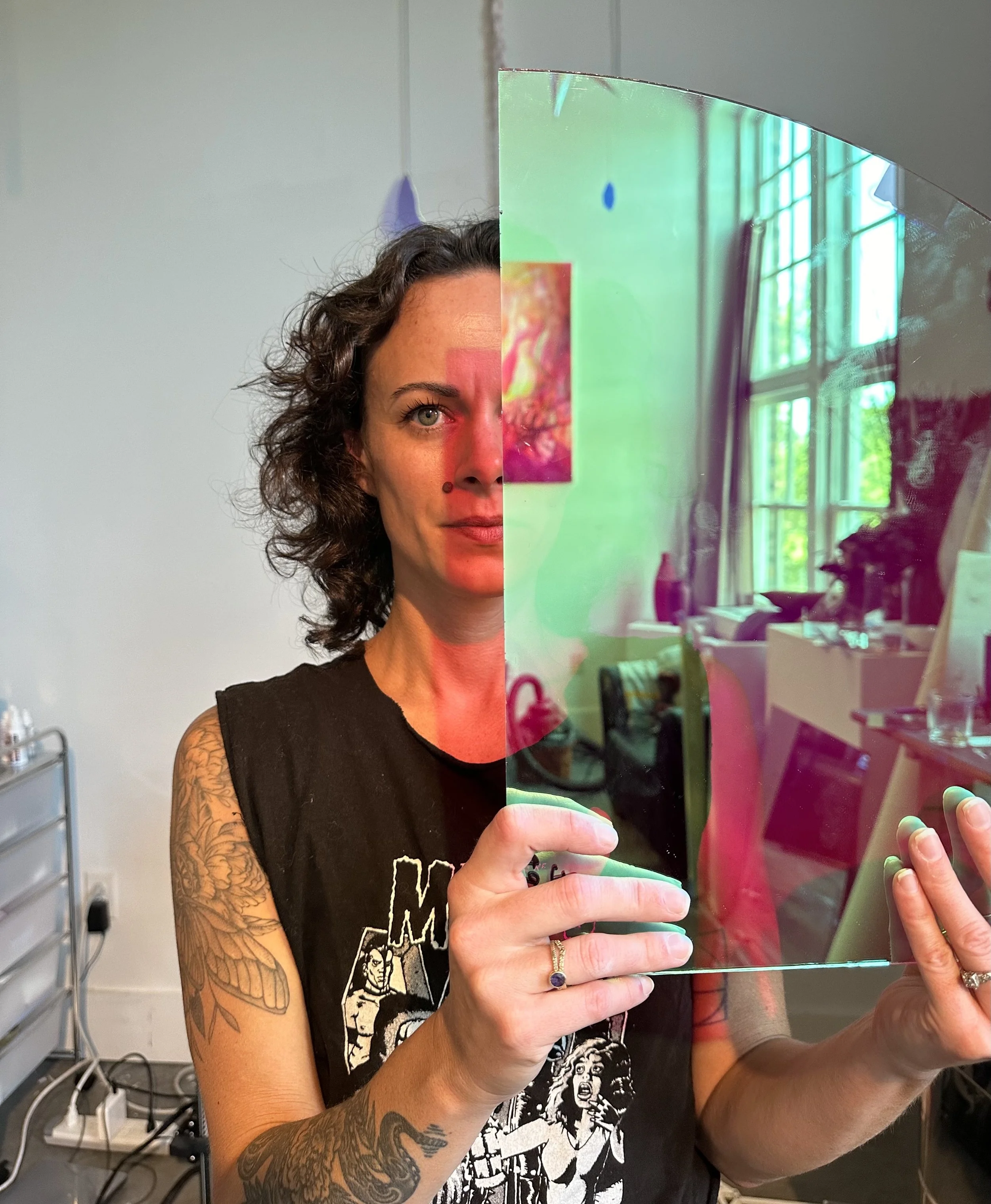 A woman with curly hair and tattoos on her arm is holding a reflective, iridescent panel in front of her face. The panel covers half of her face, revealing one eye and part of her forehead, and the reflection shows a room with large windows and greenery outside.