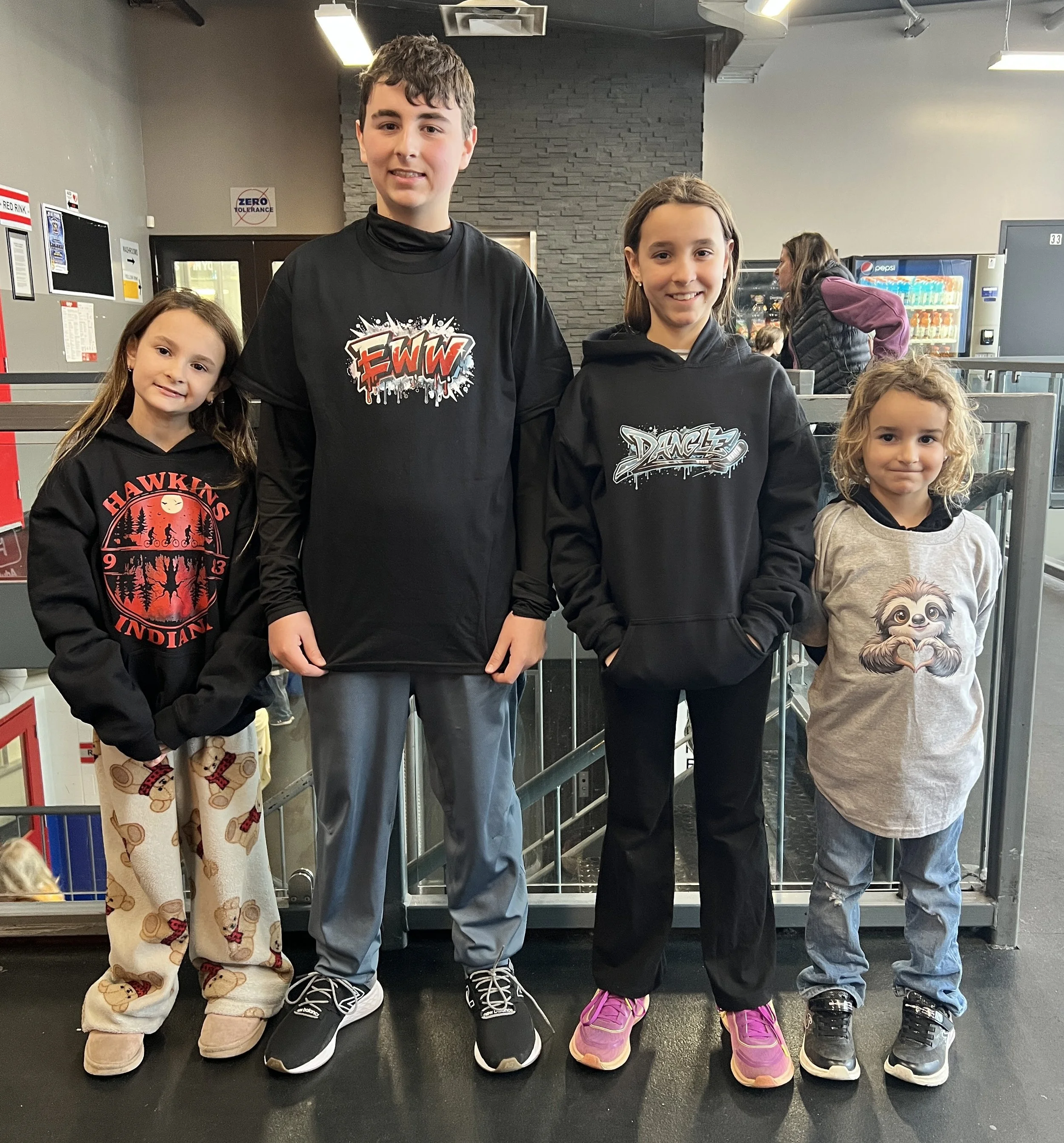 Four children standing in a row inside a building with vending machines and a gray brick wall in the background. They are wearing casual clothes, with the children in the middle wearing black hoodies that have graphics and text on them, and the girl on the left and the girl on the right in pajamas and sneakers. The children are smiling or looking at the camera.