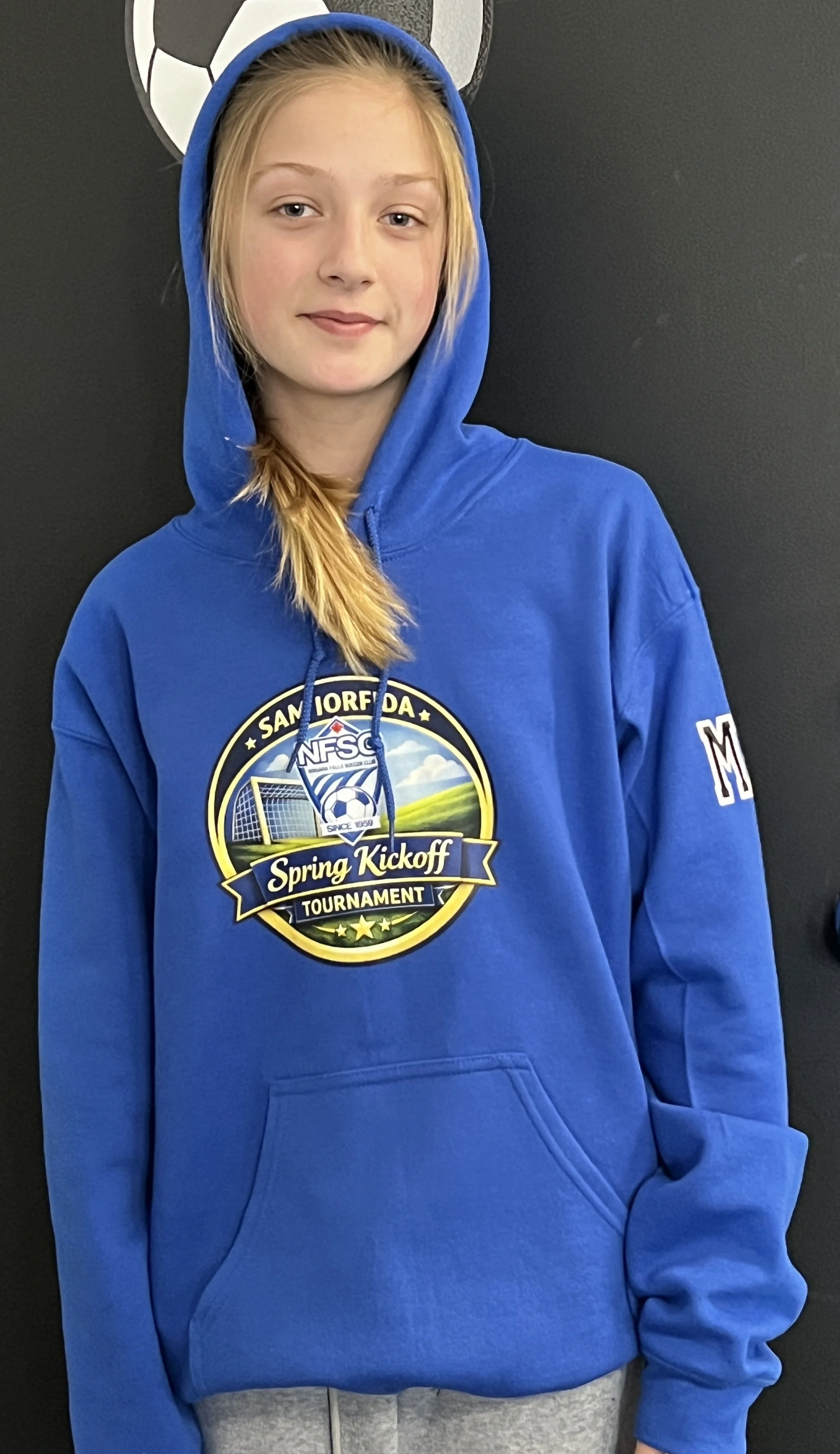 A girl wearing a blue hoodie with a soccer tournament logo, standing against a black background.