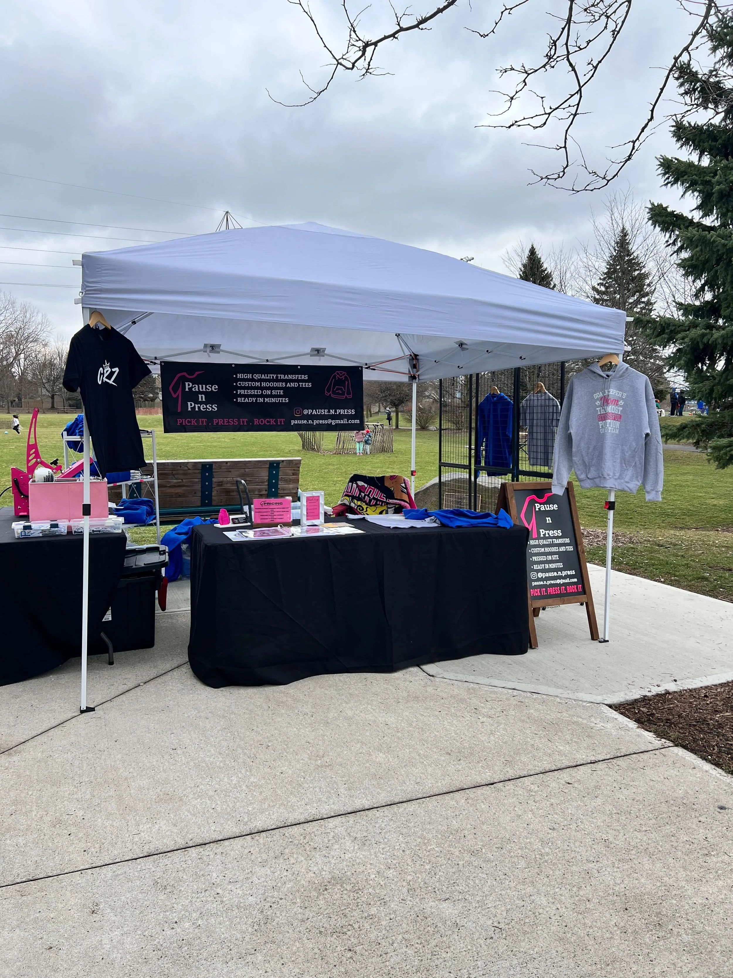 Outdoor vendor booth with clothing and accessories on display, including t-shirts and hoodies, under a white canopy in a park setting.