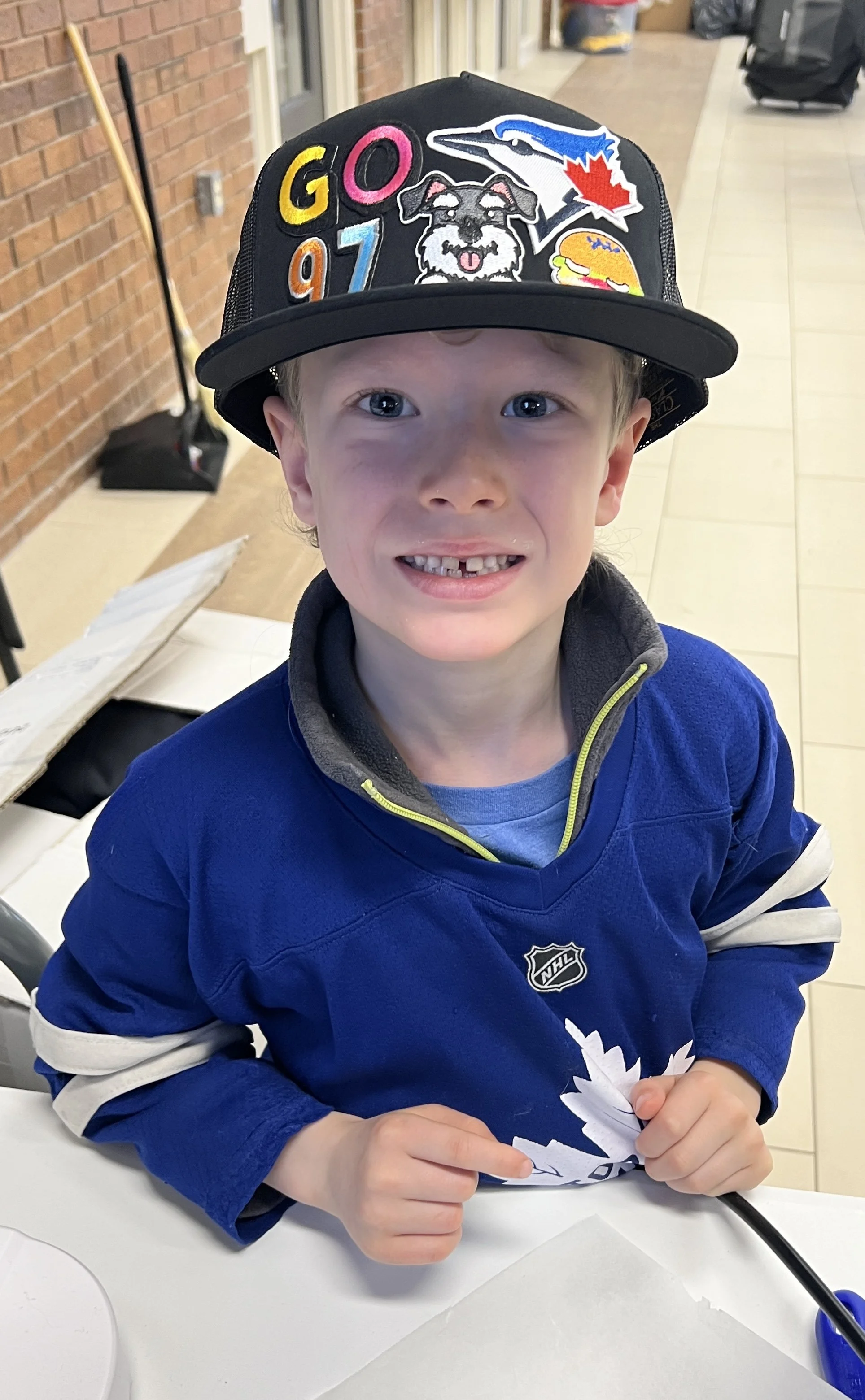A young boy with blue eyes and blonde hair smiling at the camera. He is wearing a Toronto Maple Leafs hockey jersey and a black cap with colorful patches that include a dog, a Toronto Blue Jays logo, and the numbers 97 and G.