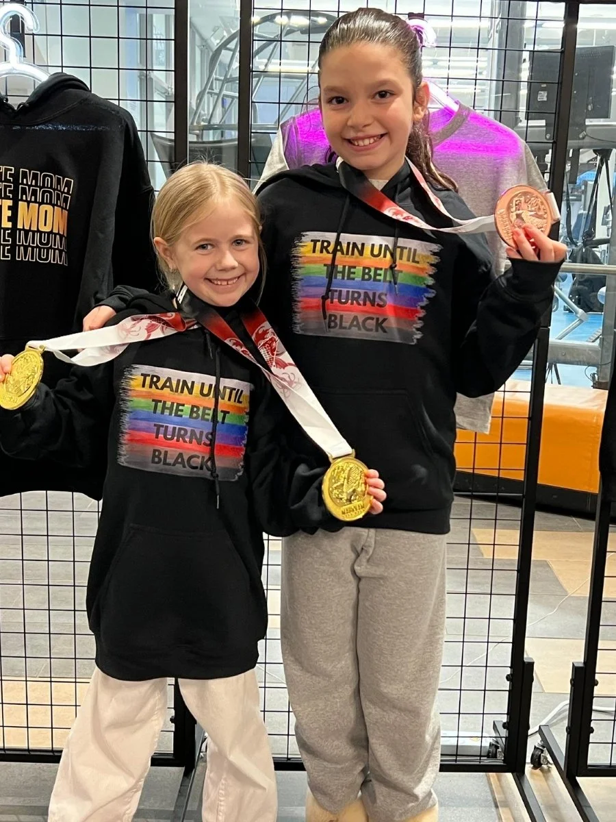 Two young girls smiling and holding medals in front of a wire fence; both are wearing black hoodies with a colorful rainbow design and the text "Train until the belt turns black." They are inside a gym or sports facility.