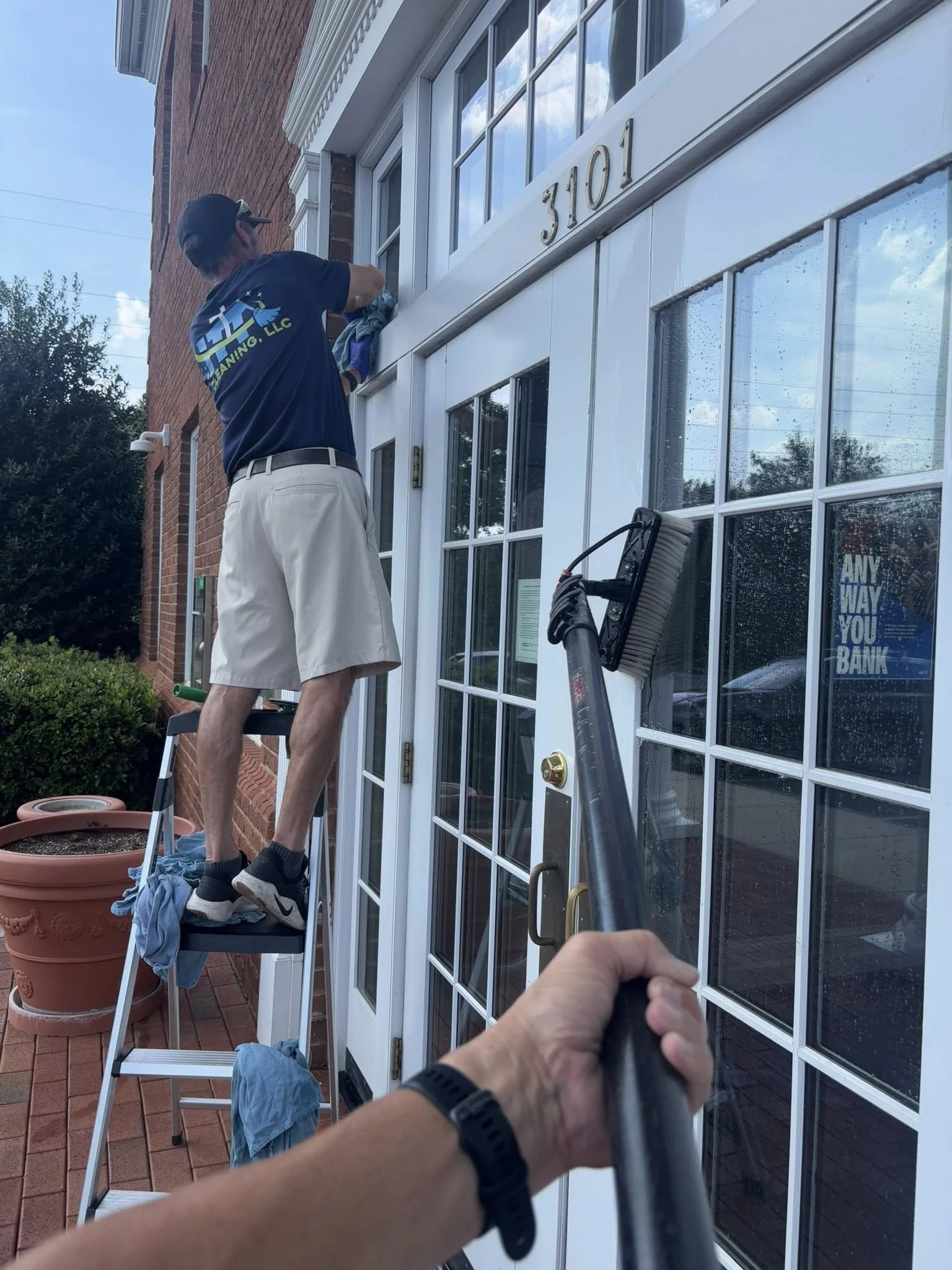 A person cleaning the exterior window of a building labeled 3101, with another person cleaning the window above, on a small ladder outside a brick building. The photo is taken from the perspective of someone holding a pressure washer or spray hose.