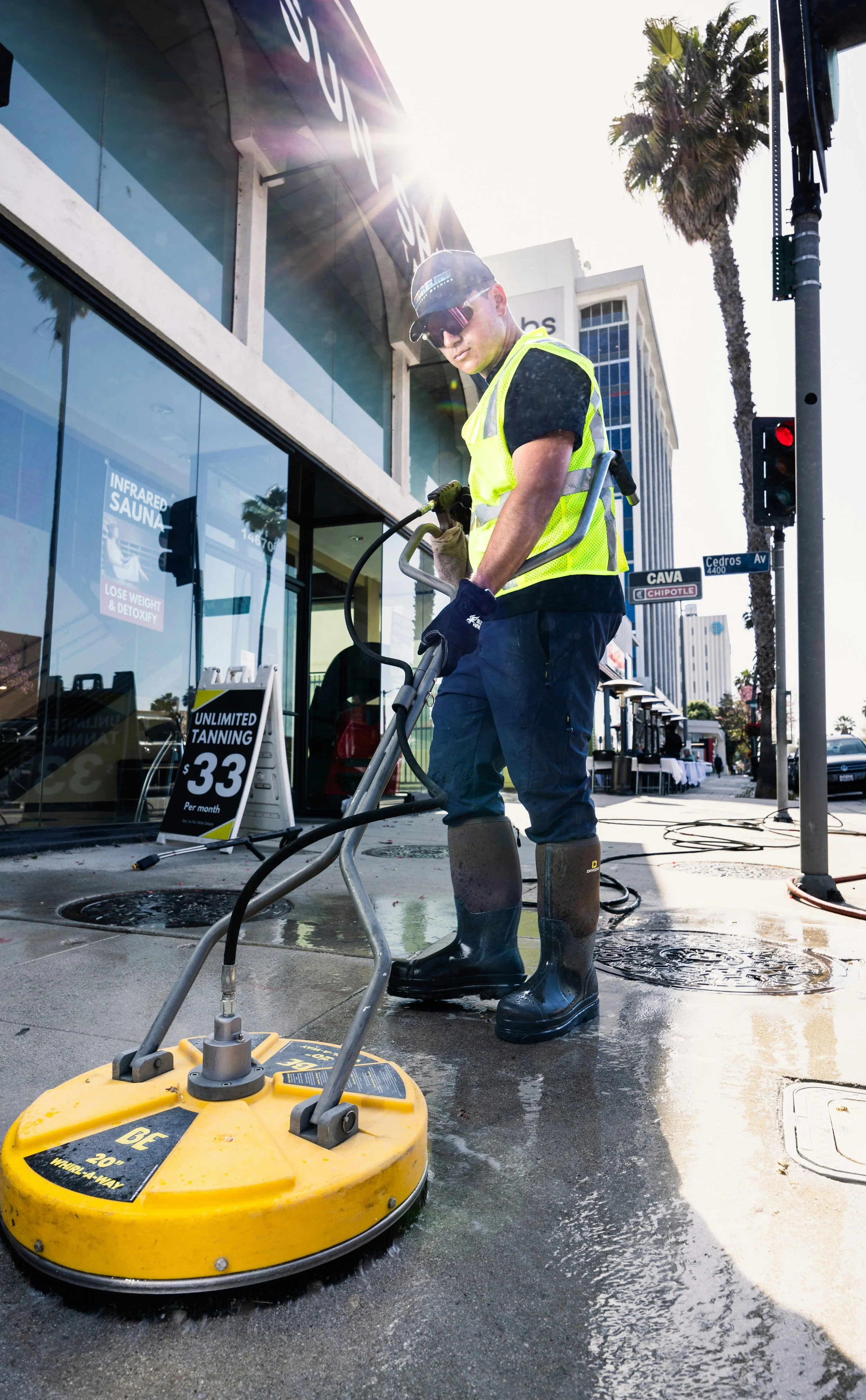 A worker in a neon yellow safety vest and rubber boots is cleaning the sidewalk with a large, yellow floor buffer. The worker is wearing sunglasses and a baseball cap, and there is sunlight shining from behind, creating a lens flare. In the background, there are street signs, palm trees, and buildings.
