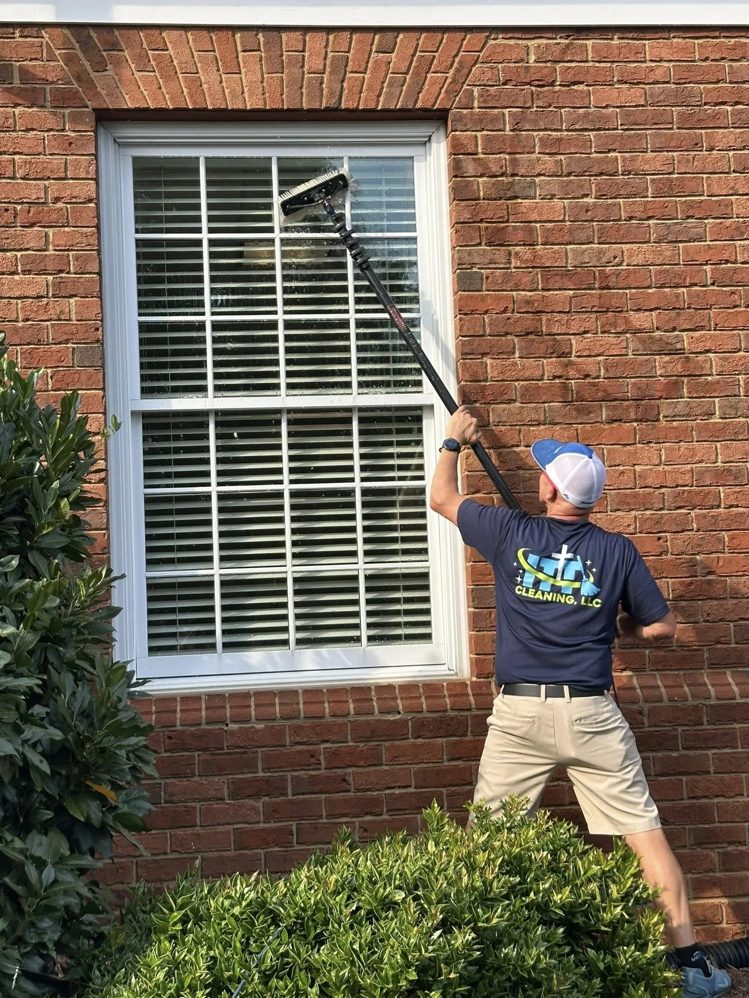 A person wearing a blue cap, blue shirt with company logo, khaki shorts, and sneakers cleaning a window with a long-handled brush, standing on a garden lawn in front of a brick house.
