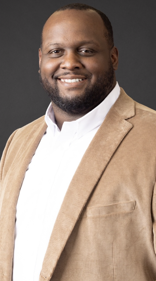 A smiling Black man wearing a beige blazer and white shirt against a dark background.