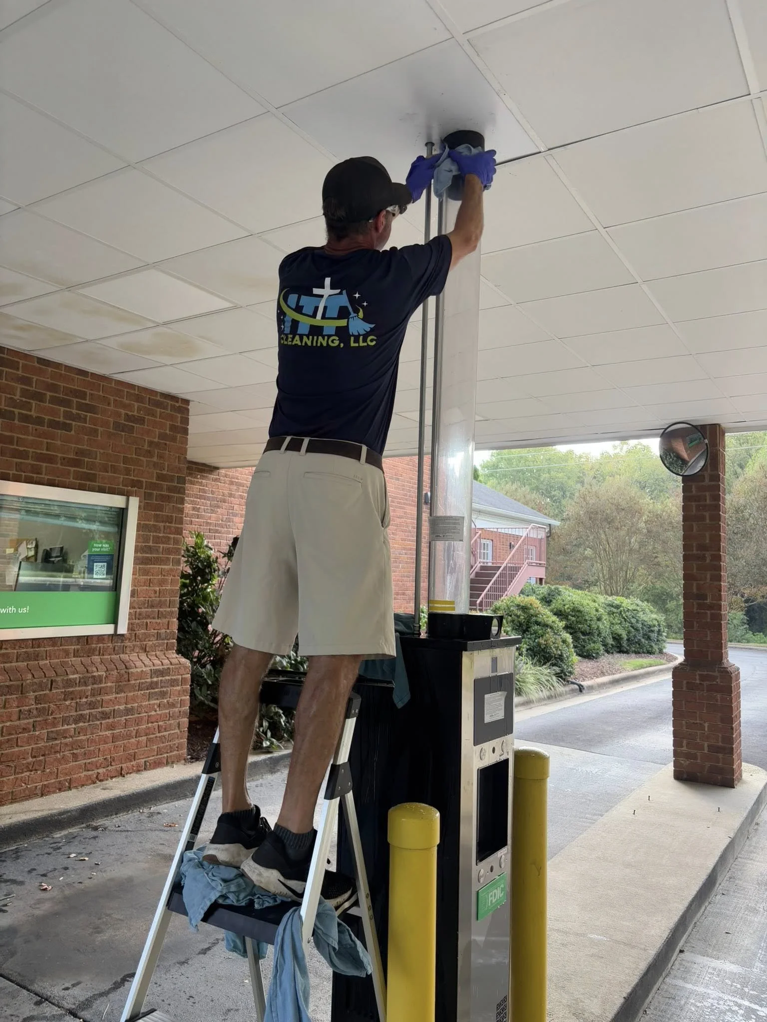 A man is cleaning the ceiling of a covered outdoor area, standing on a step ladder. He is wearing a black cap, glasses, a blue shirt with 'CLEANING LLC' on the back, and shorts. He is using a cloth and a pole to clean the ceiling tiles.