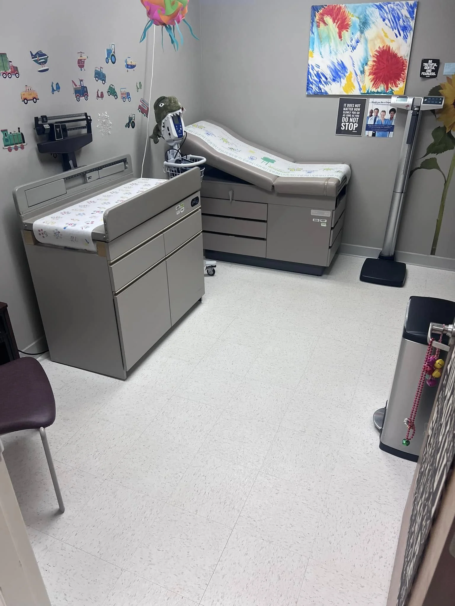 A hospital exam room with a child's examination table, a stool, a wall-mounted scale, a colorful abstract painting, and various posters and decorations, including a dinosaur plush toy and a moon balloon.