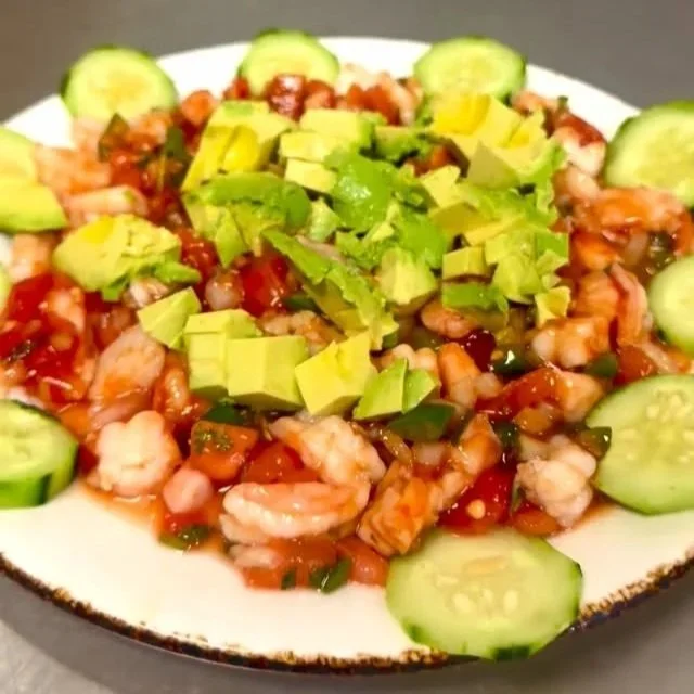 Shrimp ceviche topped with diced avocado slices, served with cucumber slices around the perimeter on a white plate