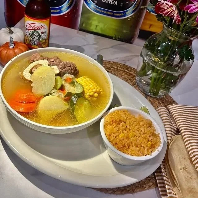 A bowl of traditional Mexican caldo with vegetables and beef, served with a side of rice, on a white tray. In the background, there are beers, a small pumpkin, a bottle of hot sauce, and a vase with pink flowers.