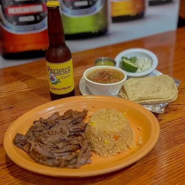 A plate of shredded beef and rice with vegetables, a small bowl of chili, a bottle of Pacifico beer, a bowl of sliced lime, chopped onion, and cilantro, and stacks of tortillas on a wooden table.
