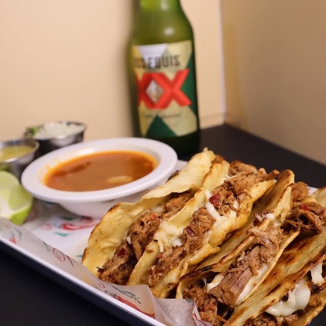 Birria tacos with shredded beef, melted cheese, lime wedge, red sauce, and a bottle of Modelo beer in the background on a black surface.