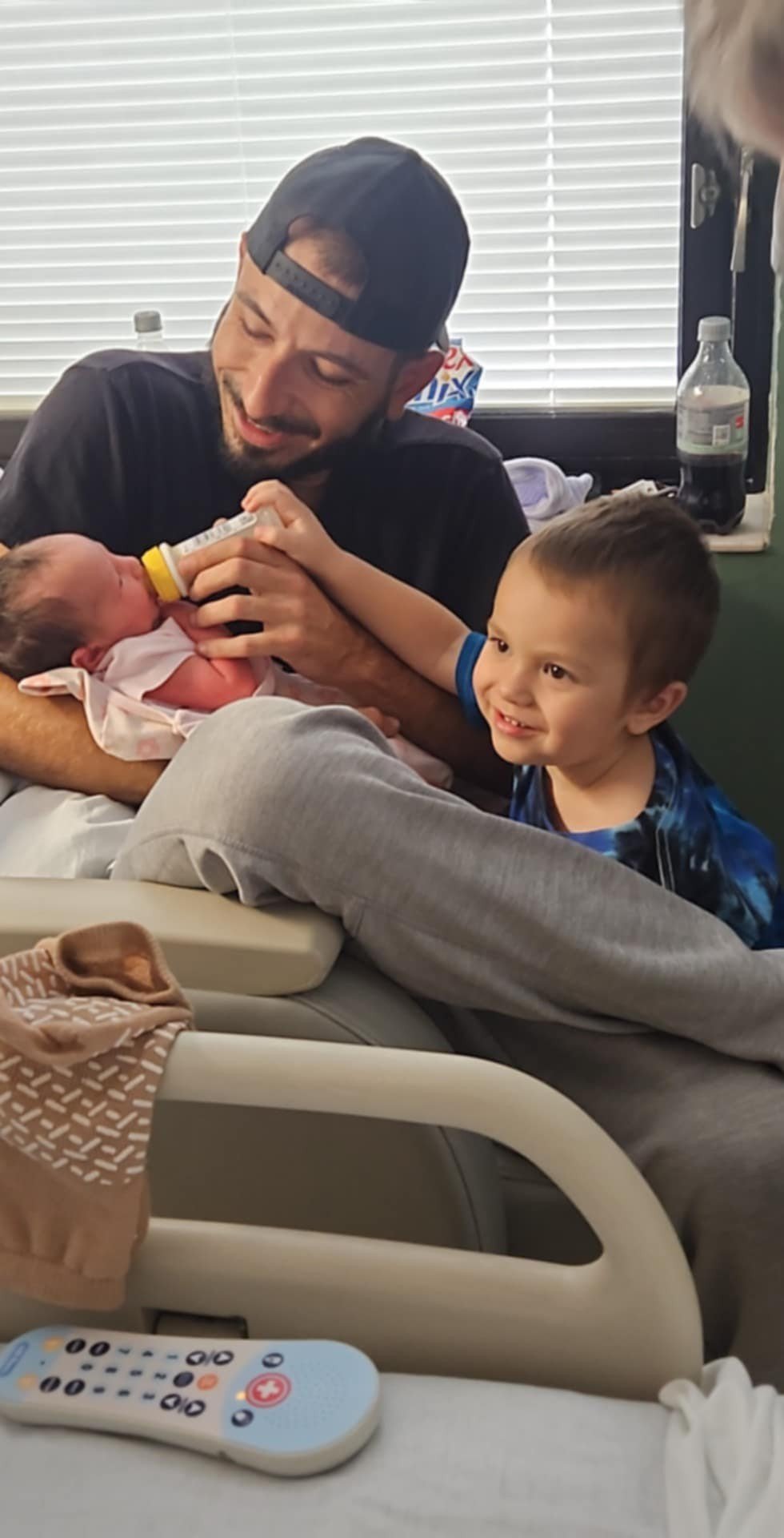A man in a baseball cap feeding a newborn baby with a bottle while a young boy watches happily in a hospital room.