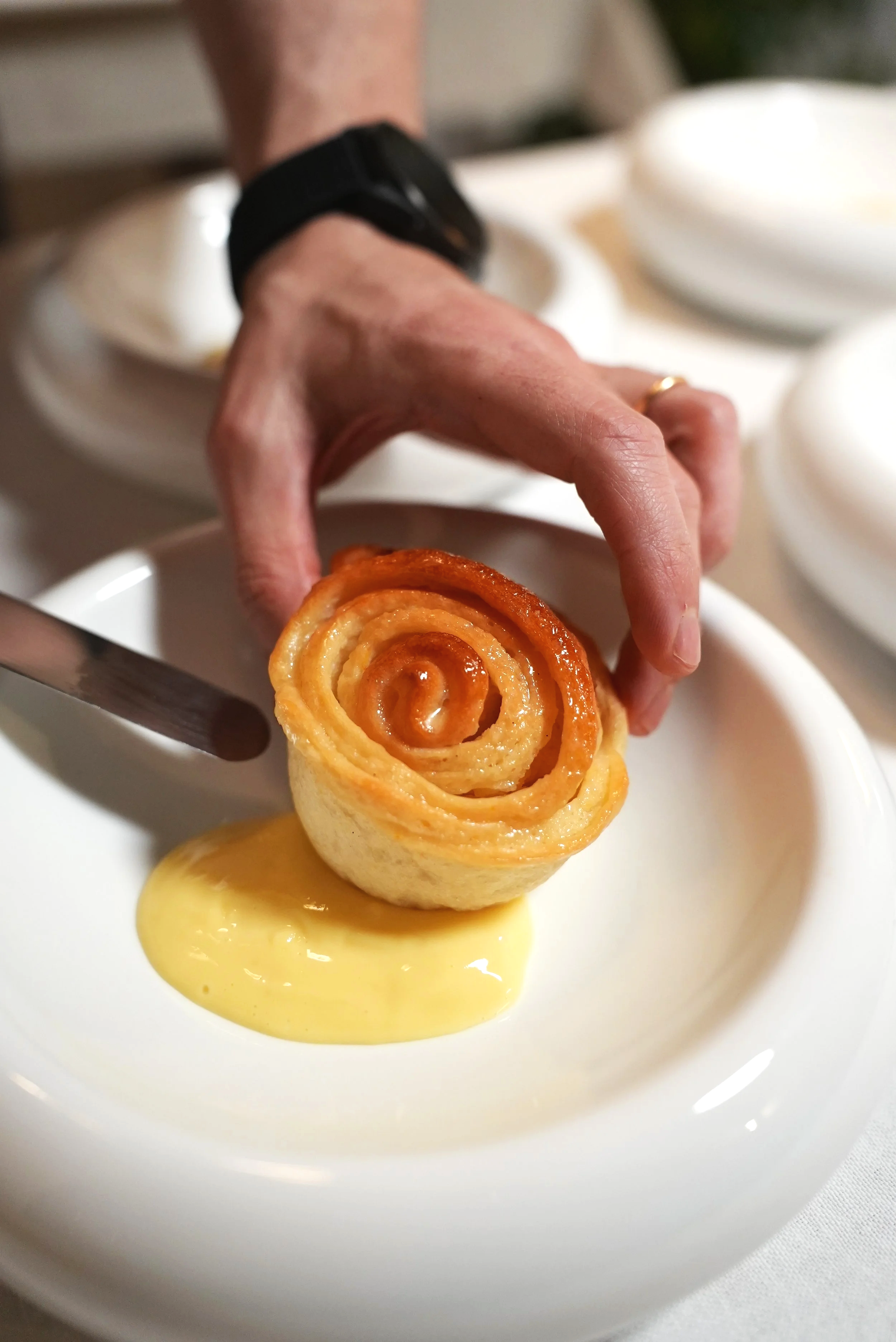 Una mano tiene in mano una corolla di pasta sfoglia ripiena, sopra un piatto bianco con una salsa gialla, con alcune ciotole di dessert in sfondo.