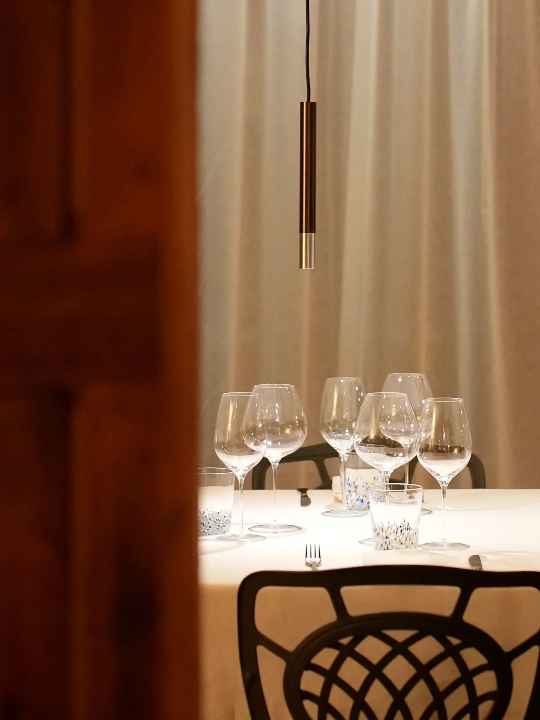 Tavolo apparecchiato con bicchieri di vino, un coltello e un cucchiaio, con cucchiai decorati, in un ambiente elegante, con sedie di design e tendaggi chiari sullo sfondo.