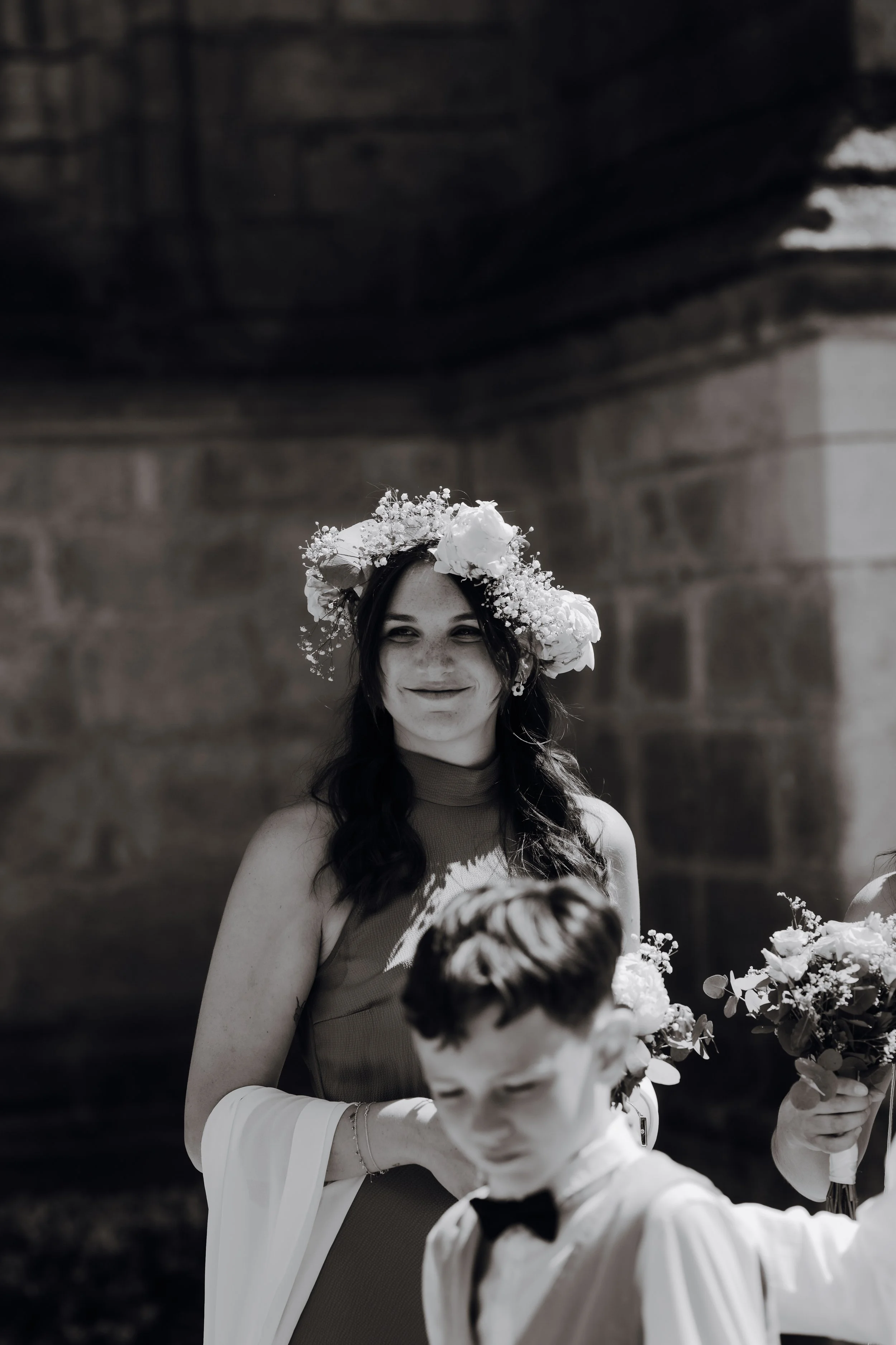 Une femme portant une couronne de fleurs et souriant lors d'une cérémonie, accompagnée d'un enfant en costume avec un nœud papillon, dans un cadre extérieur.
