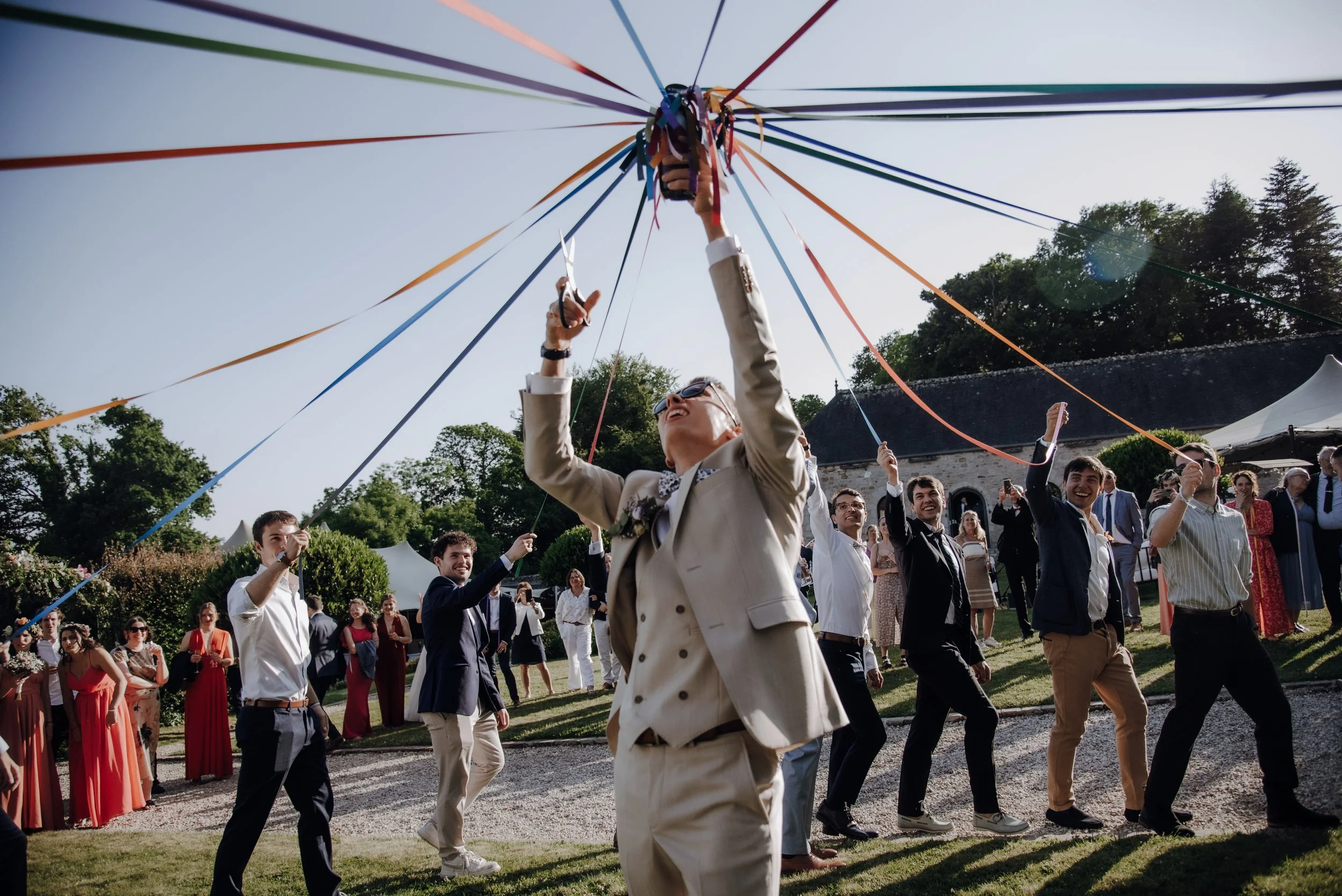 Un groupe de personnes lors d'une célébration en plein air, où des personnes dansent en cercle avec des rubans colorés attachés à un centre suspendu dans le ciel.