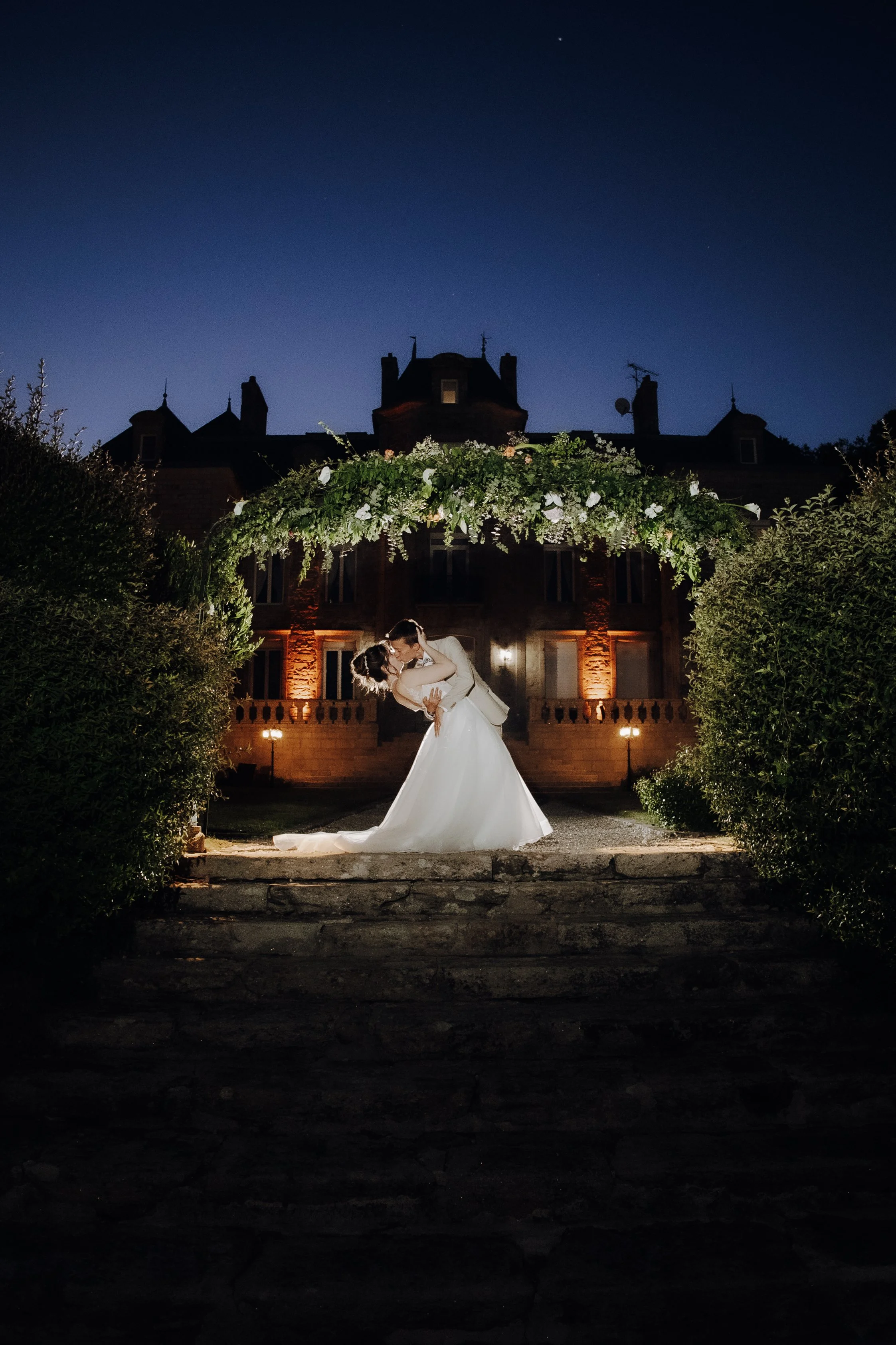 Un couple de mariés danse devant un bâtiment ancien éclairé lors d'une nuit étoilée, entouré de végétation verdoyante.