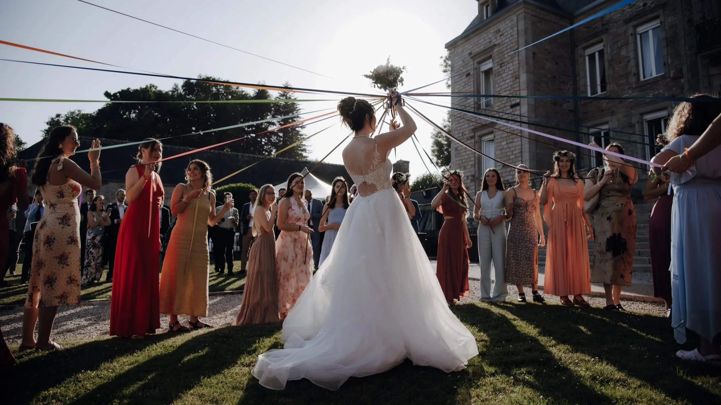 Un groupe de femmes habillées en robes longues colorées entoure une mariée en robe de mariée blanche, qui tient un bouquet en hauteur, lors d'une cérémonie en extérieur.