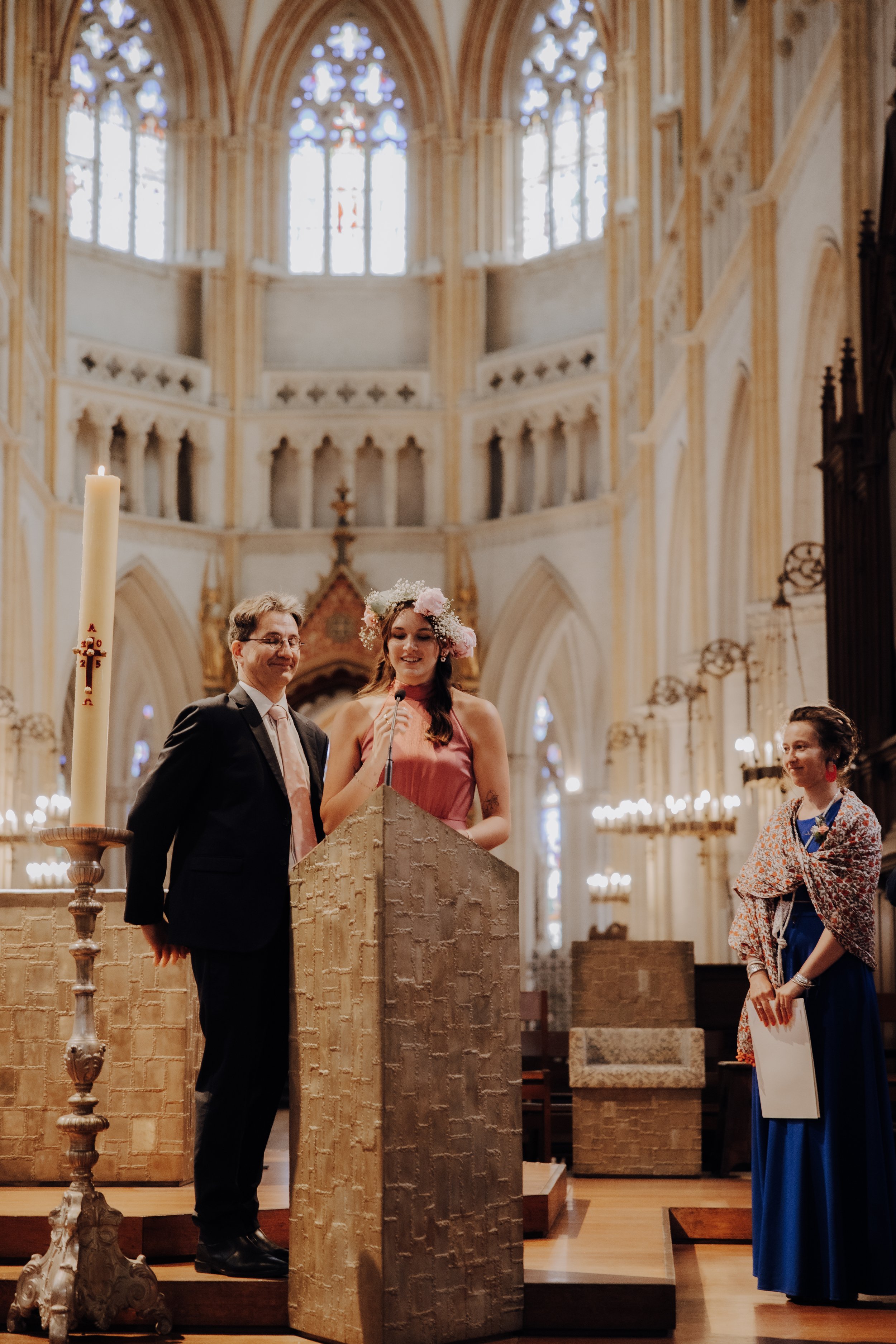 Une cérémonie religieuse dans une église avec des personnes debout, dont deux se tiennent dans une enceinte en pierre, une femme avec une couronne de fleurs et un homme en smoking, une autre femme avec un châle coloré, au fond des vitraux colorés dans une architecture gothique.