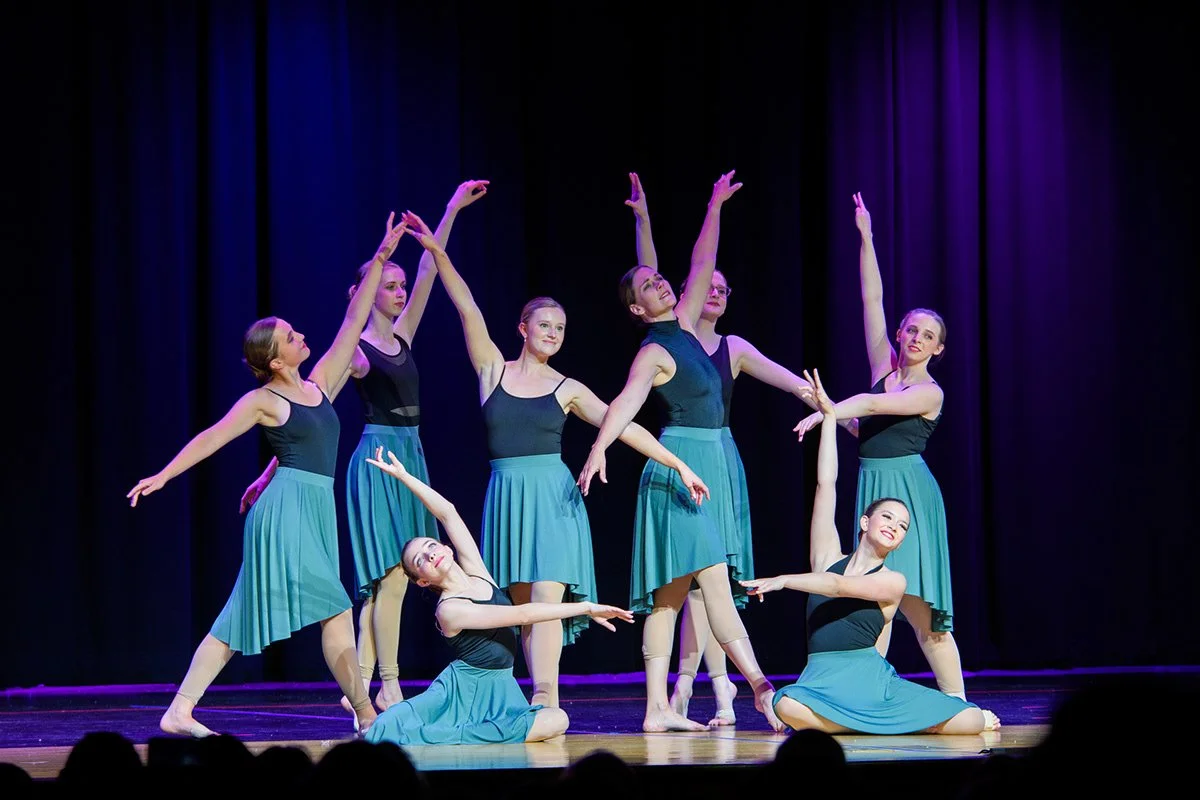 Nine ballet dancers in black and blue costumes performing on stage with purple and black curtains in the background.