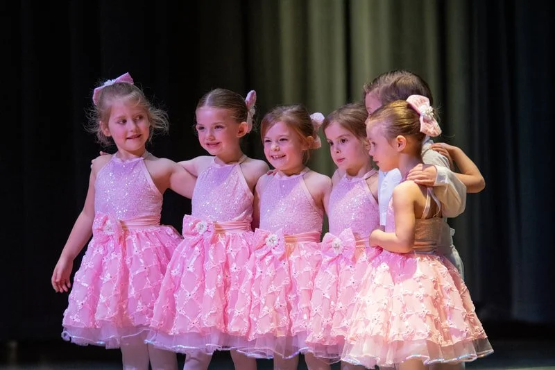 Six young girls in pink tutus and one woman in a beige outfit standing together on stage, smiling and holding each other.