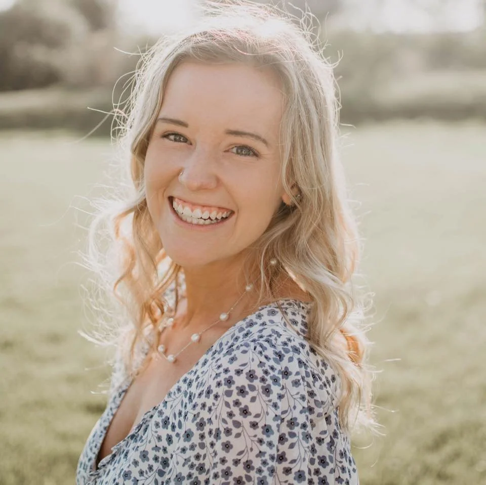 A woman with long, wavy blonde hair smiling outdoors, wearing a floral dress and a pearl necklace.