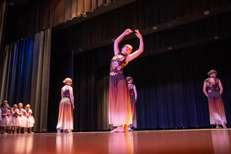 Ballet dancers performing on stage, with one dancer in front raising her arms, and a group of young dancers in pink tutus in the background.