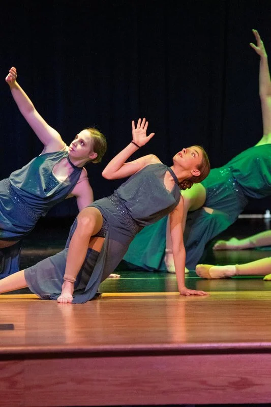 Three young women in dance costumes performing a dance on stage, with one girl in the foreground balancing on her hands and knees and others in the background also in dance poses.
