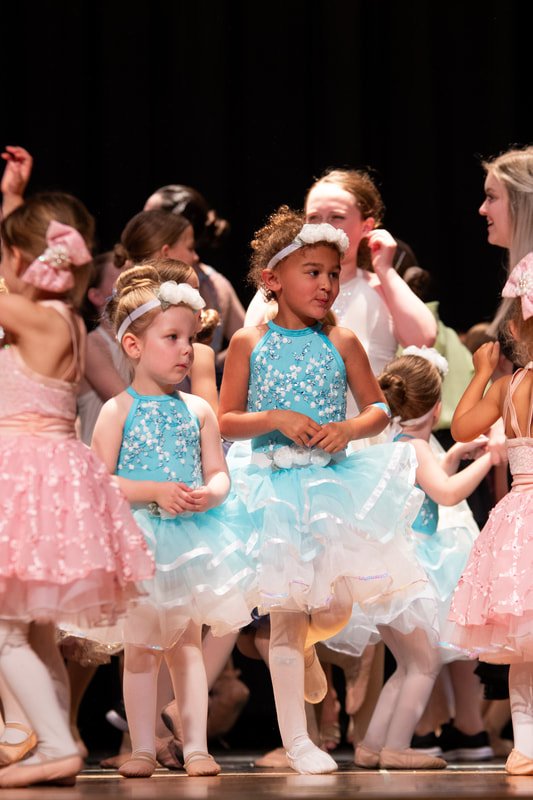 A group of young girls in ballet costumes, with some wearing blue dresses and others in pink, standing on stage.