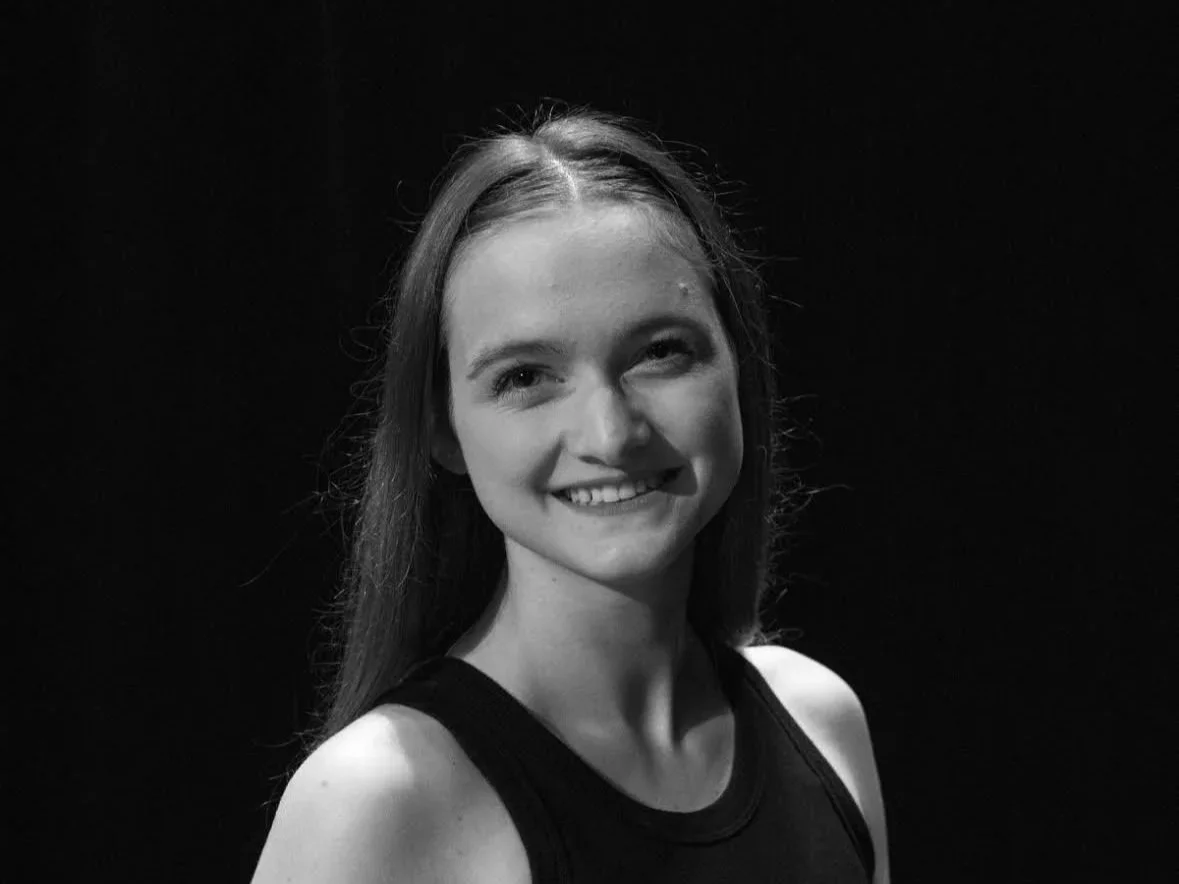 A black and white photo of a young woman with long hair, smiling, wearing a sleeveless top, against a dark background.