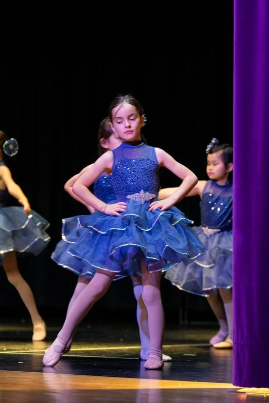 Young ballet dancer in a blue tutu with hands on hips on stage during a performance.