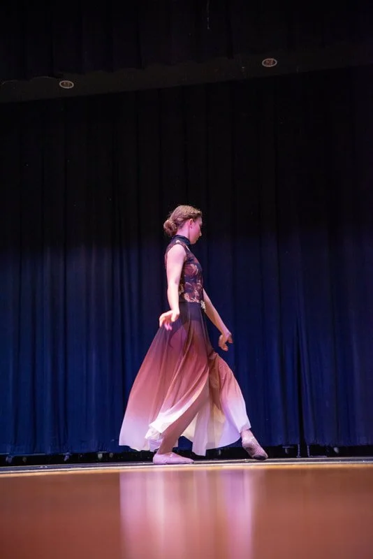 A young girl in a ballet costume practicing or performing on stage with a black curtain backdrop.