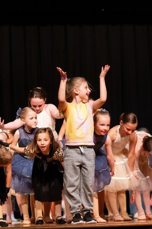 Children dressed in dance costumes on stage, bowing after a performance, with one girl in gray sweatpants and a yellow shirt standing in front, smiling and raising her hands.
