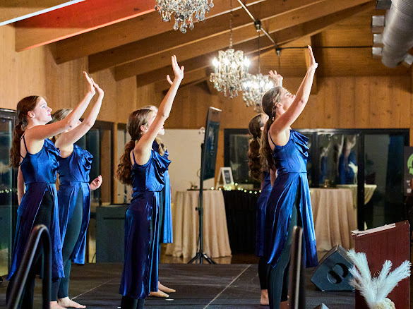 Four young girls in matching blue dresses performing a dance routine on a stage in a decorated indoor venue with chandeliers.
