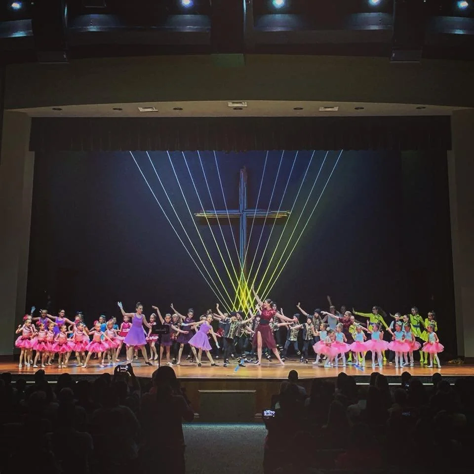 Children and adults performing a dance on stage in front of a large illuminated cross and colorful laser lights, with an audience watching.