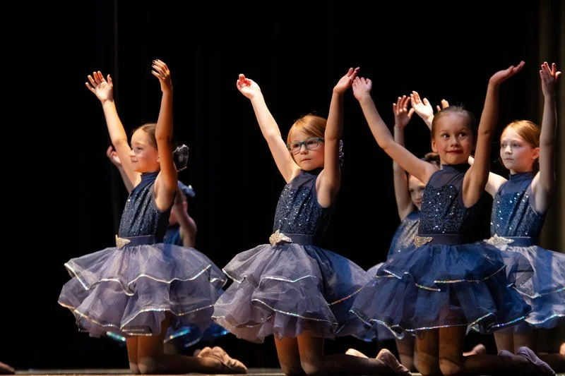 Young girls in blue glittery ballet costumes performing on stage with arms raised