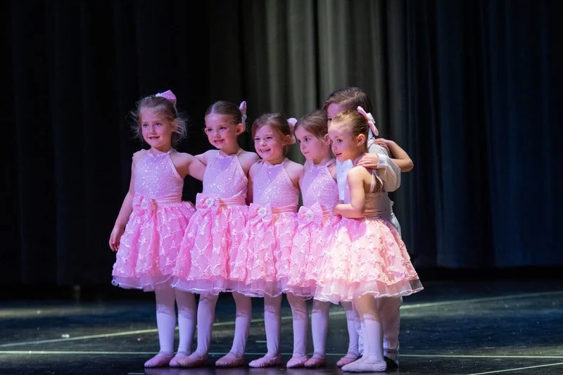 Six young girls in pink ballet costumes standing on stage and posing for a photo, with a dark curtain in the background.